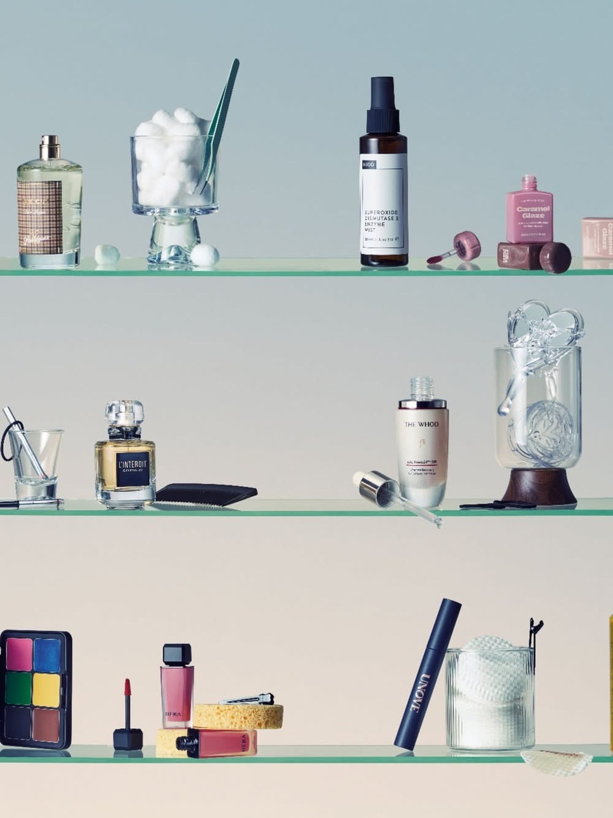 Bathroom cabinet with glass shelves holding perfume bottles, cotton pads, serums, a makeup palette and a sponge on a pale blue wall