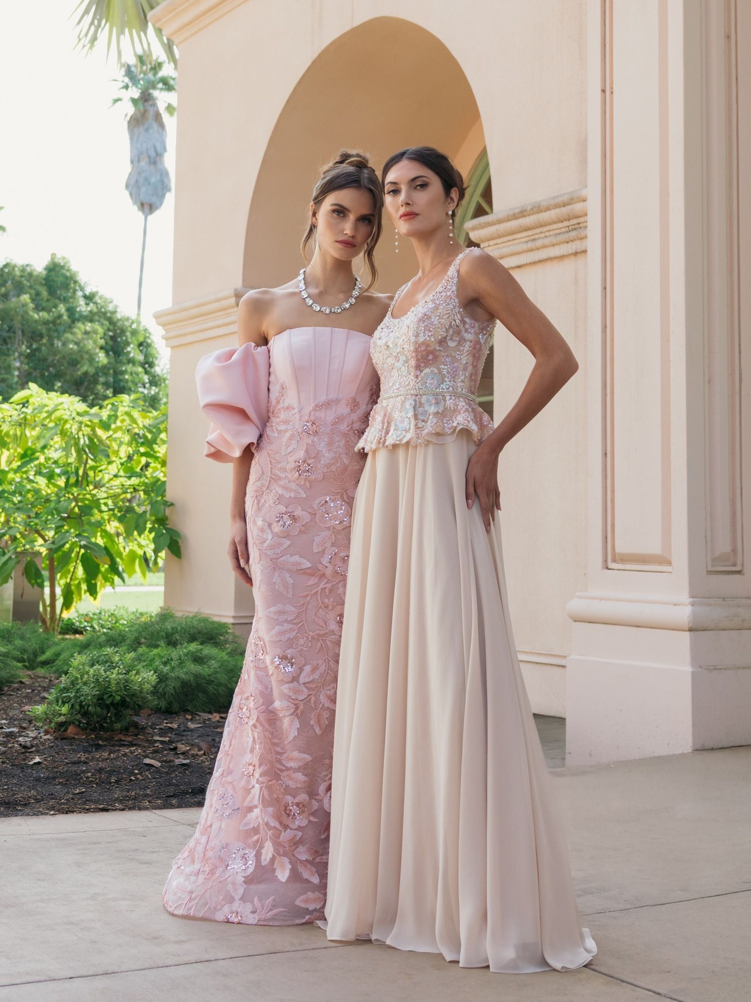 Two models in a rose appliqued gown and a champagne bustier gown in an arched courtyard
