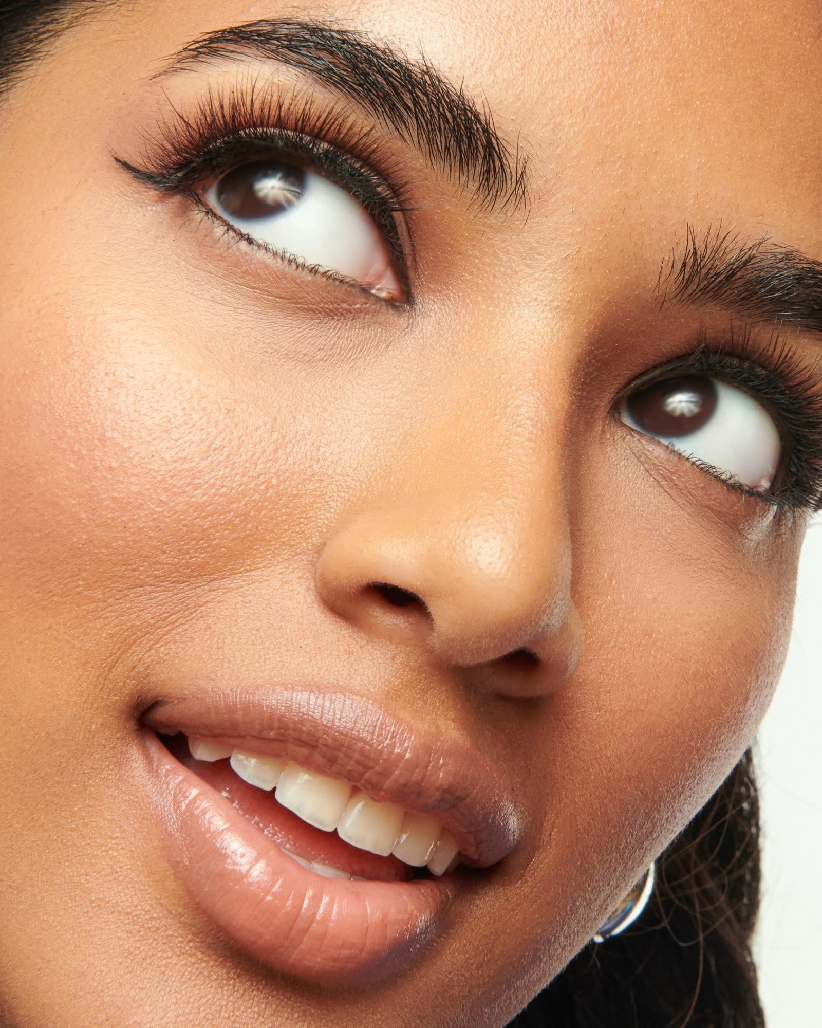 Close-up of a woman with dark brown eyes, full black lashes and a nude glossy lip, looking up and smiling