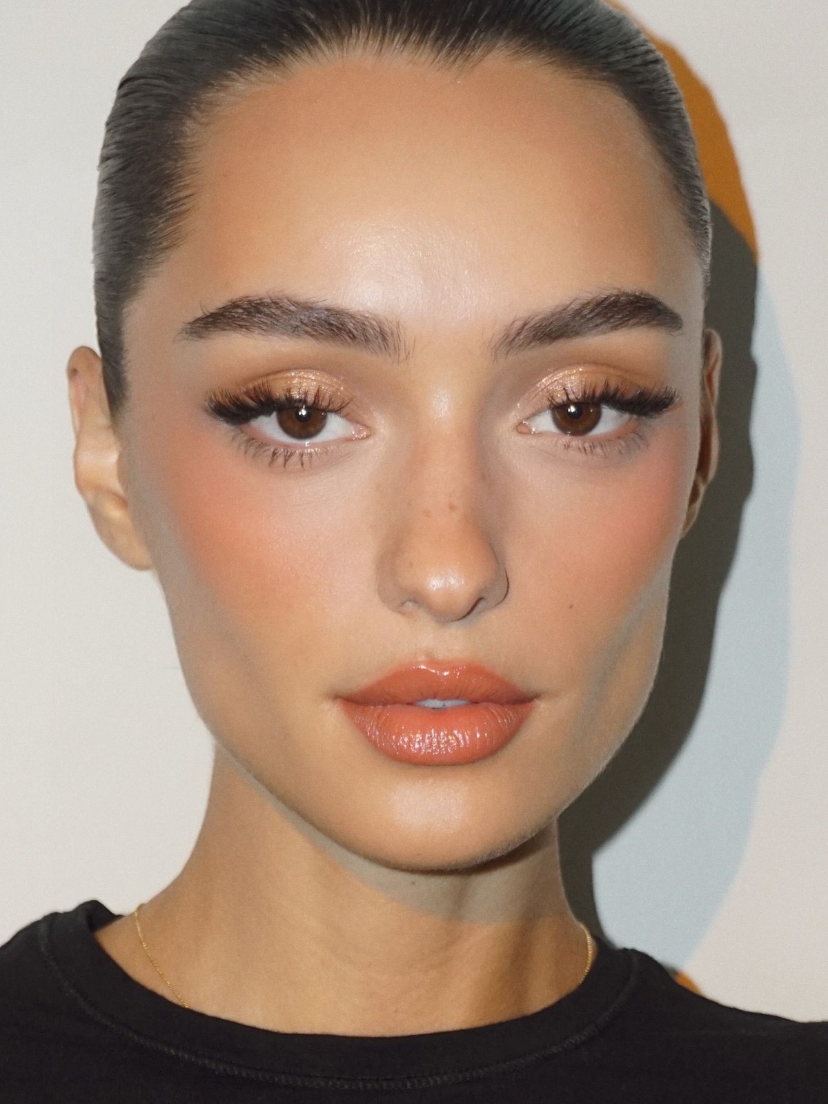 Close-up of a woman with slicked-back dark hair, peach blush, shimmery eyeshadow, long lashes and a glossy orange-toned lip
