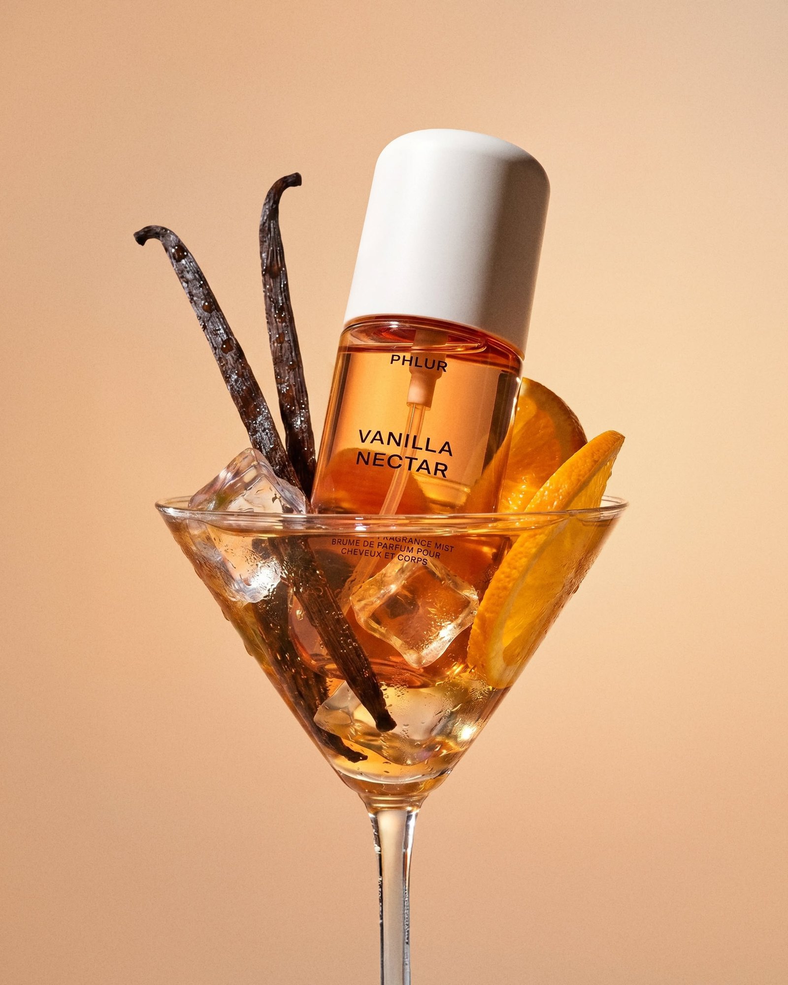 PHLUR Vanilla Nectar body mist balanced in a martini glass with vanilla pods, ice cubes and orange slices on a peach background