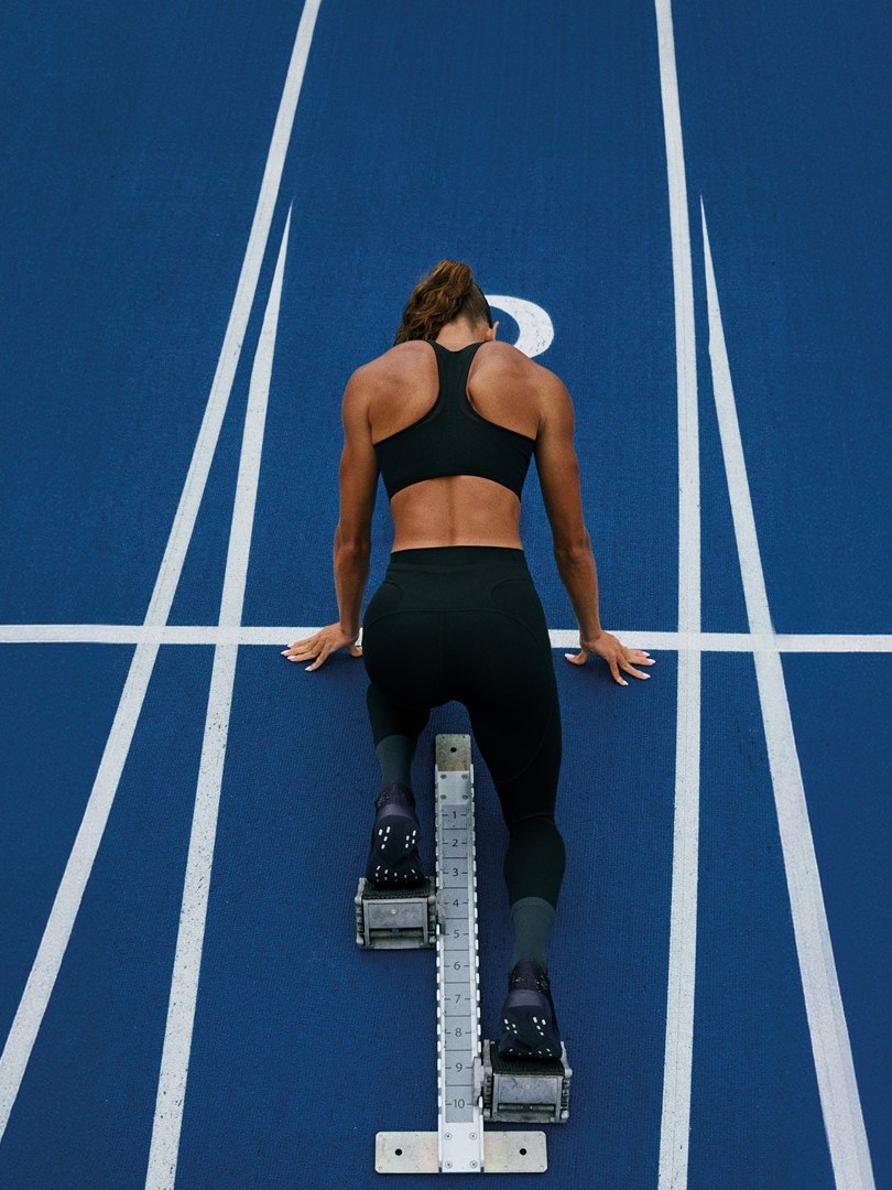 Anna Hall on a blue running track in a performance crop and leggings, viewed from behind