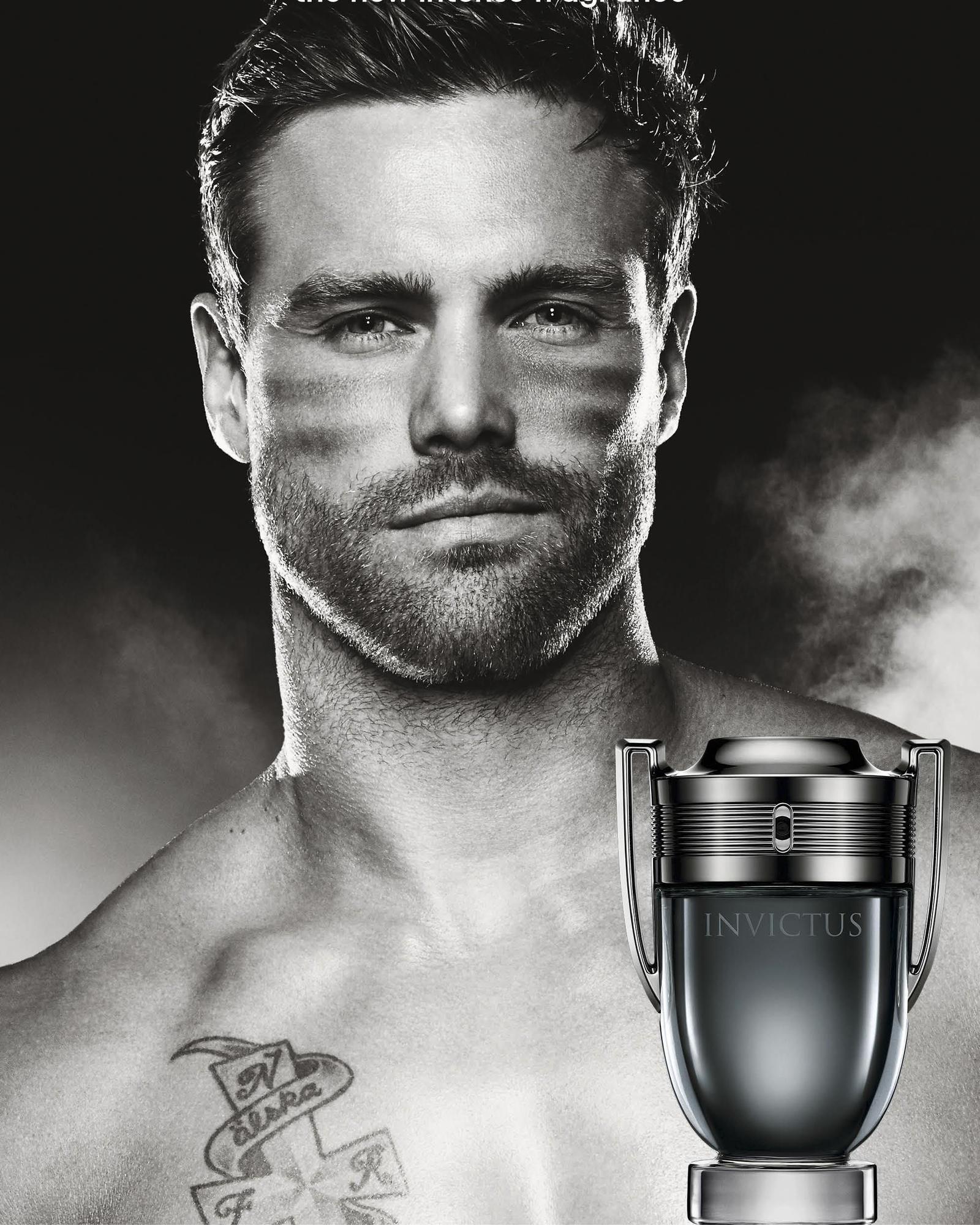 Black and white close-up portrait of a bearded man with a dark, smoky background and a trophy-shaped Invictus bottle at lower right