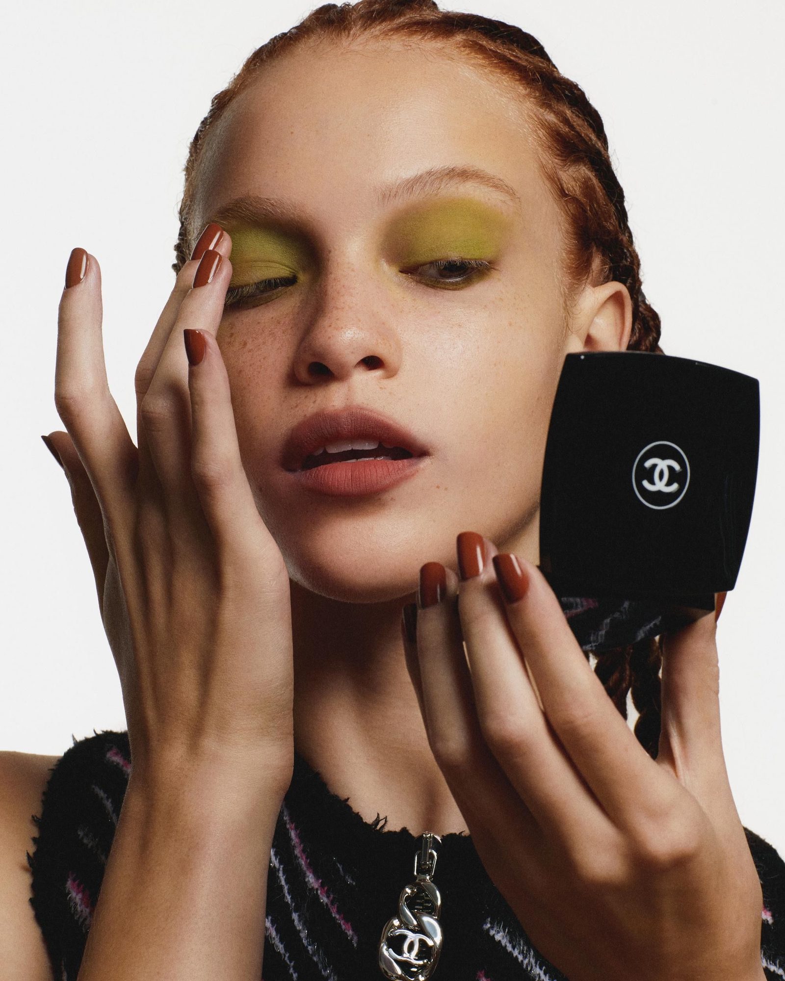 A freckled young woman with braided hair, yellow-green eyeshadow and brown-red nails, touching her eyelid with one hand while holding a black Chanel compact in the other, wearing a black tweed top with a silver chain necklace.
