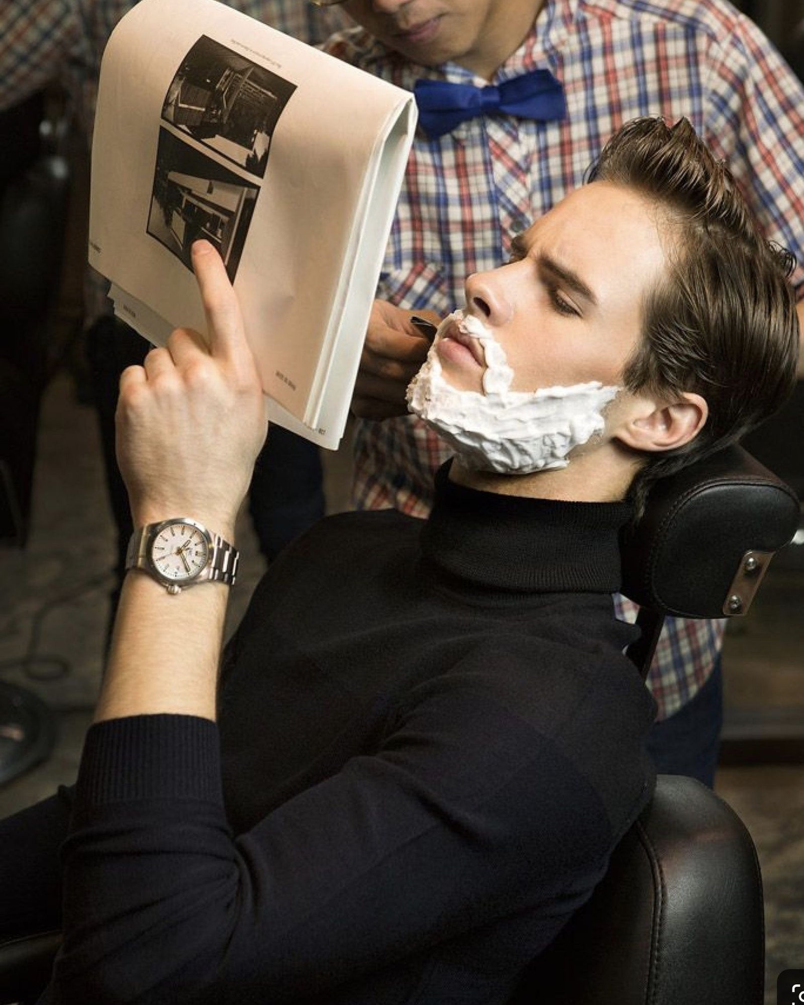Man with brown hair leaning back in a barber chair in a black turtleneck, his lower face covered in shaving foam while a barber in a plaid shirt and blue bow tie works beside him