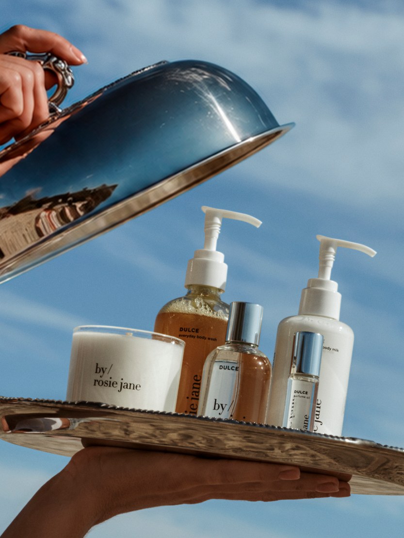 Hands lifting a silver cloche above a tray of By Rosie Jane body-care and perfume bottles