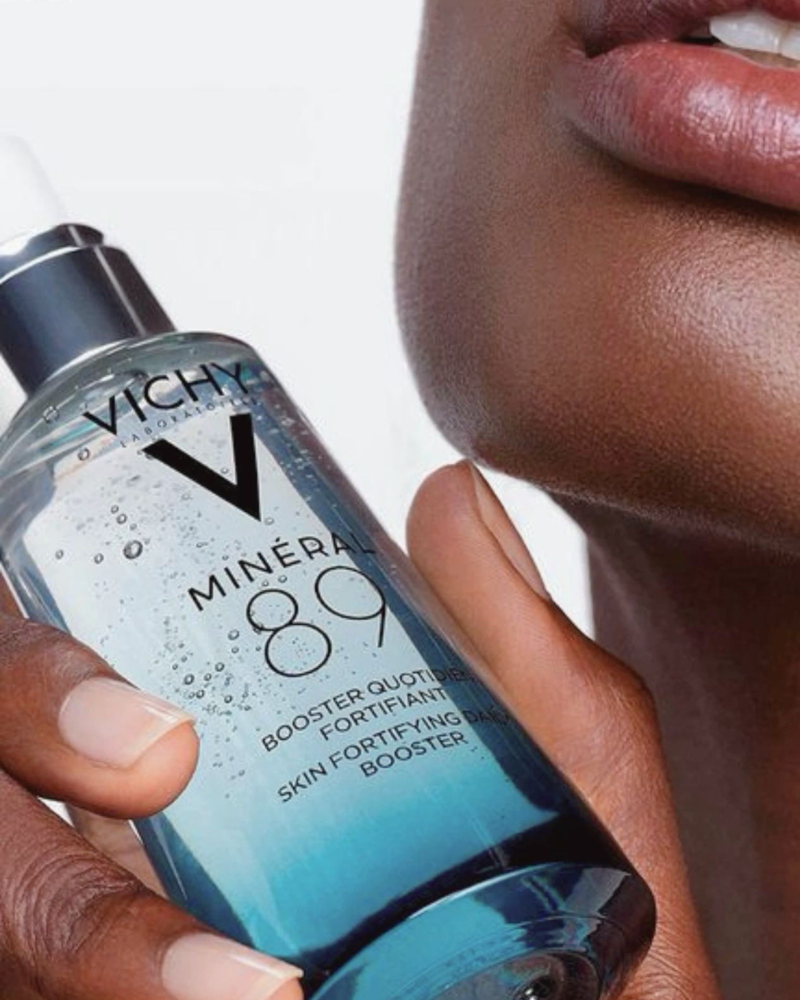 Close-up of a hand holding a Vichy Mineral 89 skin fortifying daily booster bottle beside a chin and lips