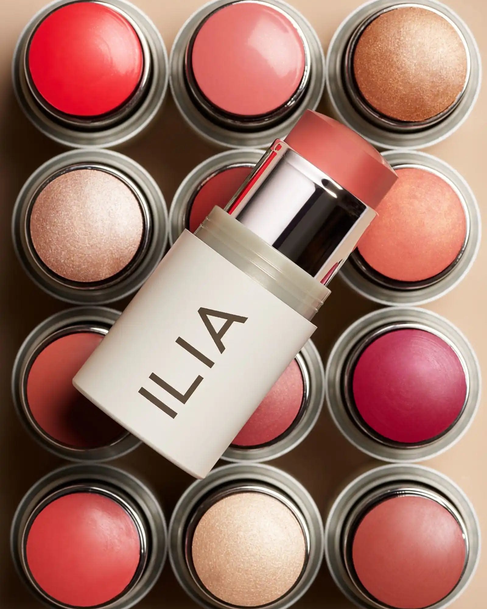 Open ILIA Multi-Stick in a pink shade lying over a grid of cream blush and highlighter tubs in corals, pinks and golds